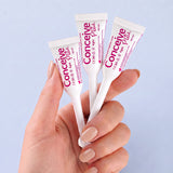 A hand with light pink manicured nails holds three white concealer tubes with pink and purple branding against a soft light blue background. The tubes are labeled 'Conceine Plus' and '12m'.